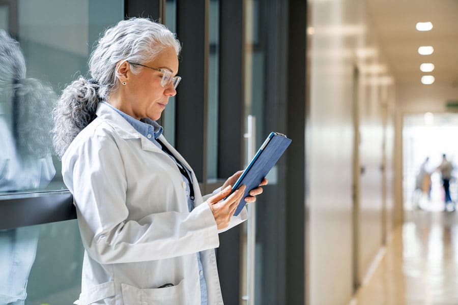 A woman with gray hair, wearing glasses and a white lab coat, stands in a Sterling Heights hallway and looks at a tablet. She appears to be an MI interventional pain management professional reviewing information.