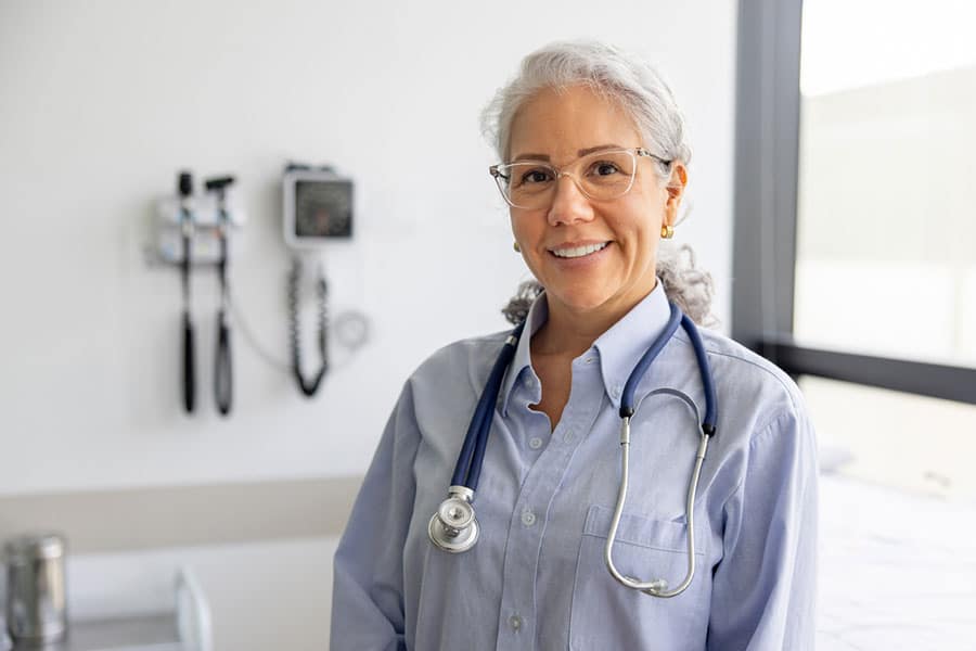 A smiling doctor with gray hair and glasses, specializing in Interventional Pain Management, stands in a bright Sterling Heights, MI examination room with equipment on the wall behind her.