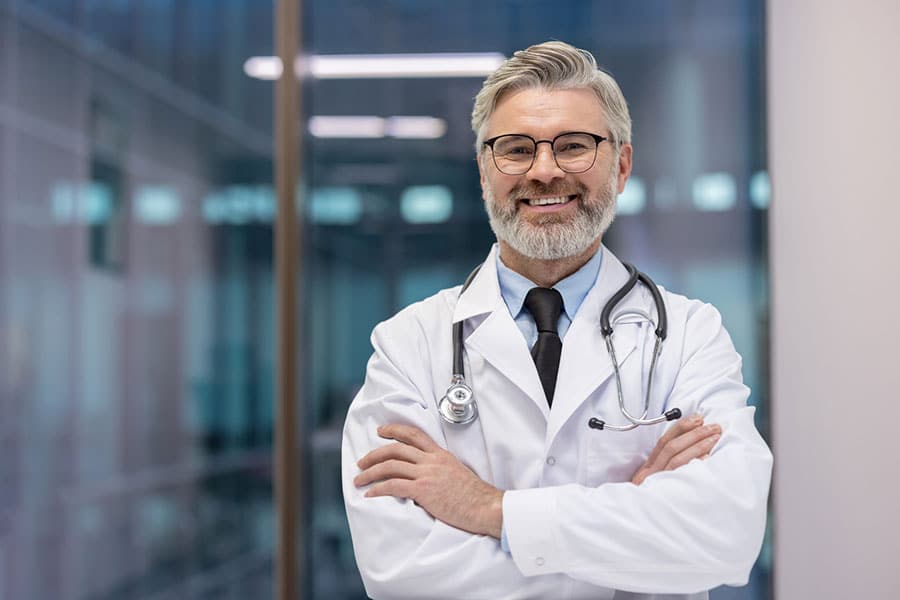 A smiling male doctor specializing in Interventional Pain Management, with gray hair and beard, wearing glasses, a white lab coat, and stethoscope, stands with arms crossed in a modern Sterling Heights, MI medical facility.
