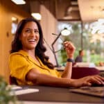 A woman in a yellow shirt sits at a desk with a laptop, smiling and holding her glasses—showing no signs of hand pain—against the backdrop of a modern office with large windows and bright lighting.