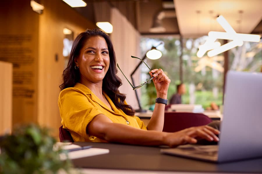 A woman in a yellow shirt sits at a desk with a laptop, smiling and holding her glasses—showing no signs of hand pain—against the backdrop of a modern office with large windows and bright lighting.