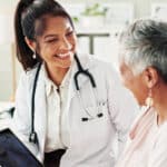 A smiling doctor with a stethoscope shows a tablet to an older woman, discussing non-surgical joint pain management. They are both seated, engaged in a friendly conversation in a bright medical office.