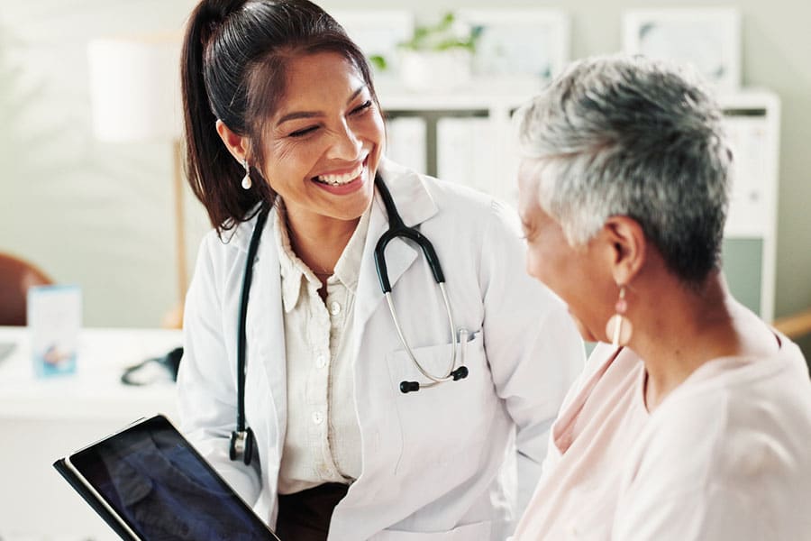 A smiling doctor with a stethoscope shows a tablet to an older woman, discussing non-surgical joint pain management. They are both seated, engaged in a friendly conversation in a bright medical office.