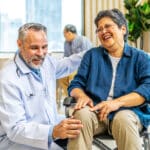 A smiling doctor in a white coat gently holds the knee of a laughing woman in a wheelchair, highlighting compassionate knee pain treatment. Indoors, others converse in the background, creating a warm, friendly atmosphere.
