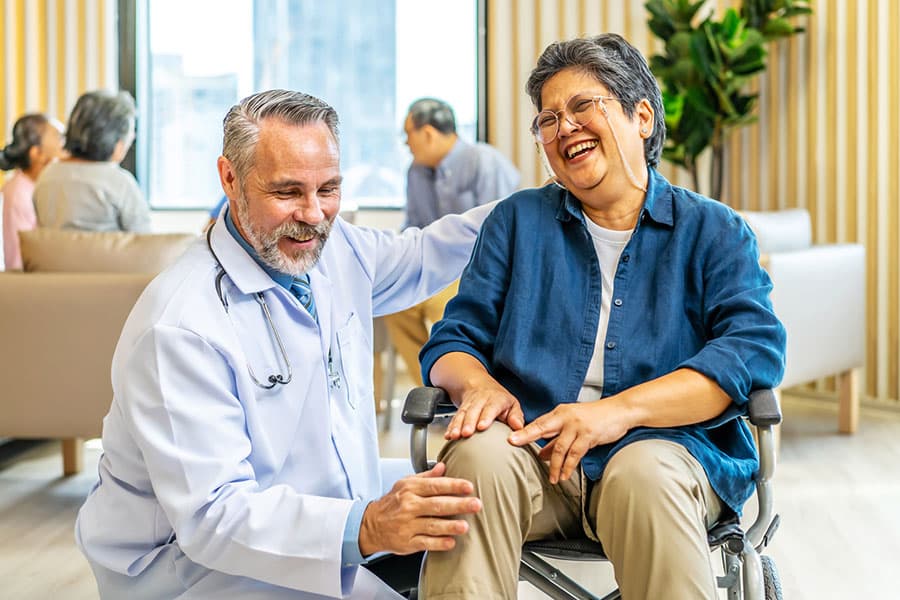 A smiling doctor in a white coat gently holds the knee of a laughing woman in a wheelchair, highlighting compassionate knee pain treatment. Indoors, others converse in the background, creating a warm, friendly atmosphere.