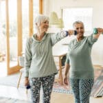 Two older women in comfortable workout clothes lift small dumbbells and smile while improving mobility together in a bright, cozy living room with wooden floors and large windows.