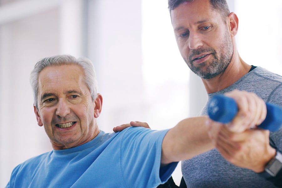 An older man in a blue shirt lifts a small dumbbell with his arm extended, while a trainer in gray helps guide him through physical therapy focused on maintaining mobility and improving movement during the exercise session.