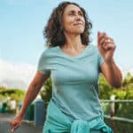 A middle-aged woman with curly hair, enjoying a brisk walk on a sunny day, shows how staying active can help manage hand pain from carpal tunnel without surgery. She wears a light blue T-shirt and has a jacket tied around her waist.