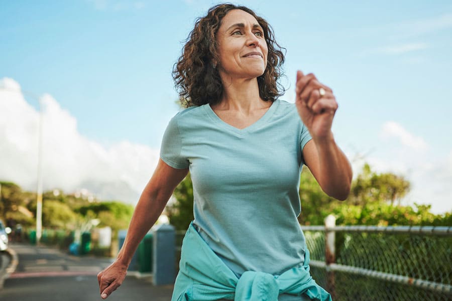 A middle-aged woman with curly hair, enjoying a brisk walk on a sunny day, shows how staying active can help manage hand pain from carpal tunnel without surgery. She wears a light blue T-shirt and has a jacket tied around her waist.