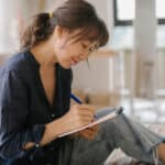 A woman sits on a couch, smiling and writing in a notebook with a blue pen. She appears focused as she works on her care plan, surrounded by bright natural light streaming through large windows.