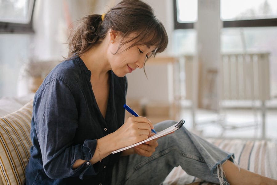 A woman sits on a couch, smiling and writing in a notebook with a blue pen. She appears focused as she works on her care plan, surrounded by bright natural light streaming through large windows.