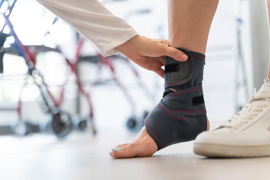 A healthcare professional fits a black ankle brace with red trim on a person's foot to help manage ankle pain. The person, possibly from Macomb County, wears a white sneaker on the other foot, and a walker is visible in the blurred background. A healthcare professional fits a black ankle brace with red trim on a person's foot to help manage ankle pain. The person, possibly from Macomb County, wears a white sneaker on the other foot, and a walker is visible in the blurred background.