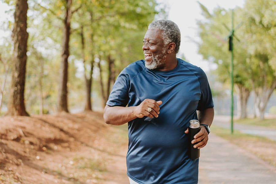 Older man in a blue shirt jogging outdoors on a tree-lined path in Dearborn MI, smiling and holding a water bottle, with sunlight filtering through the trees.