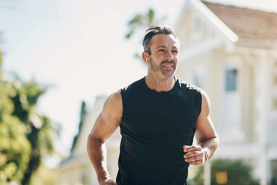 A man in a sleeveless black shirt jogs outdoors on a sunny day in Farmington Hills, MI, with trees and a house blurred in the background. He appears focused and is wearing earphones.