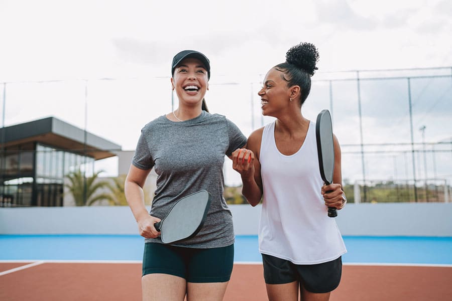 Two women smiling and walking arm-in-arm on an outdoor pickleball court in Macomb County, each holding a paddle, dressed in athletic wear, enjoying a sunny day—staying active and mindful of ankle pain and Achilles tendon injuries. Two women smiling and walking arm-in-arm on an outdoor pickleball court in Macomb County, each holding a paddle, dressed in athletic wear, enjoying a sunny day—staying active and mindful of ankle pain and Achilles tendon injuries.