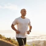 A man in a white shirt and black shorts jogs outdoors on a hill at sunrise, smiling, with a scenic view of Macomb County’s city and water in the background—enjoying his run without ankle pain or Achilles tendon injuries.