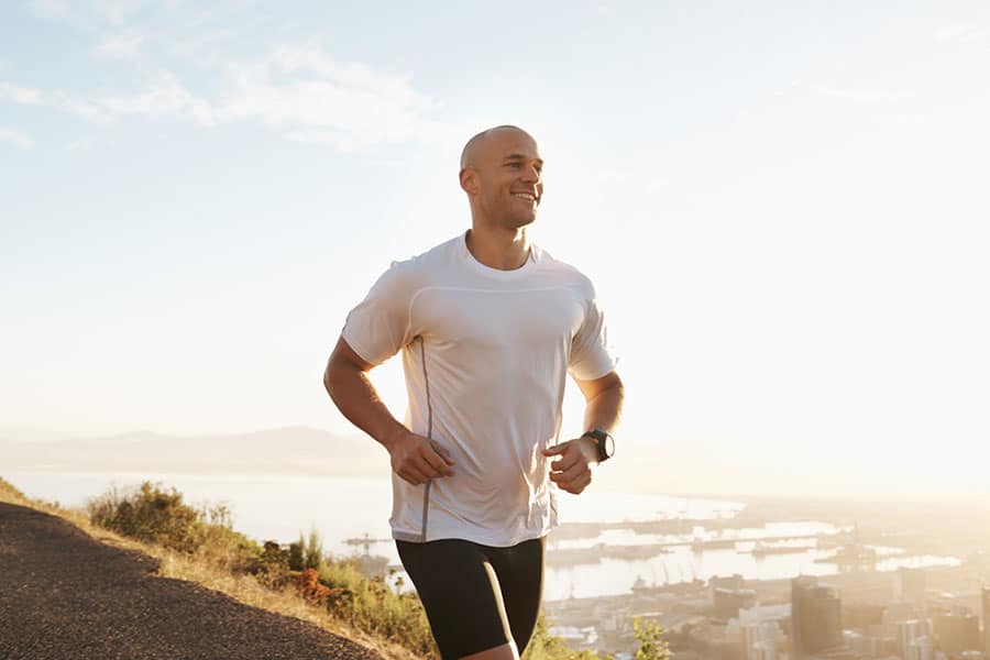 A man in a white shirt and black shorts jogs outdoors on a hill at sunrise, smiling, with a scenic view of Macomb County’s city and water in the background—enjoying his run without ankle pain or Achilles tendon injuries.