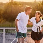 A man and a woman stand on a tennis court in Macomb County, smiling and laughing together at sunset. Both hold tennis rackets and wear sporty outfits, enjoying the game while staying mindful of avoiding ankle pain or Achilles tendon injuries.
