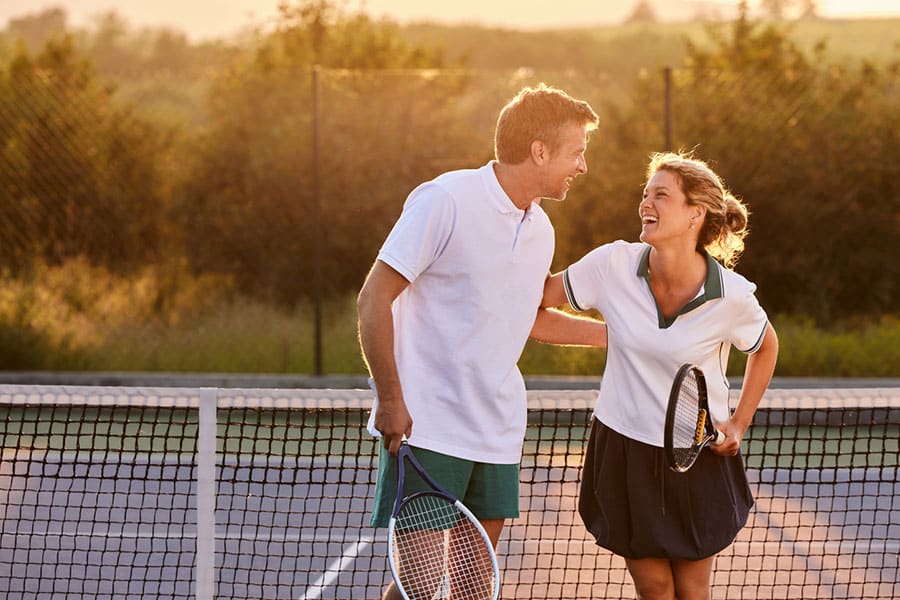 A man and a woman stand on a tennis court in Macomb County, smiling and laughing together at sunset. Both hold tennis rackets and wear sporty outfits, enjoying the game while staying mindful of avoiding ankle pain or Achilles tendon injuries.