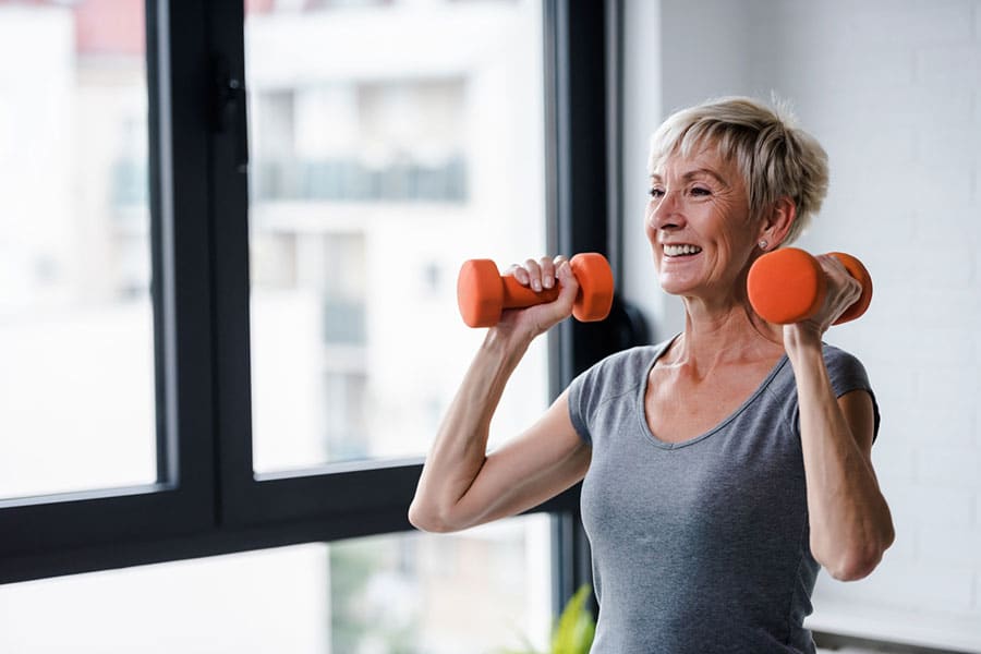 Smiling older woman with short gray hair lifts orange dumbbells indoors near a window in Wayne County, wearing a gray shirt and appearing focused and energetic—an inspiring example of active living and joint pain treatment.
