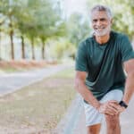 Smiling older man in athletic wear stretches outdoors on a tree-lined path, resting his hands on one knee to help relieve low back pain, with greenery and sunlight in the background.