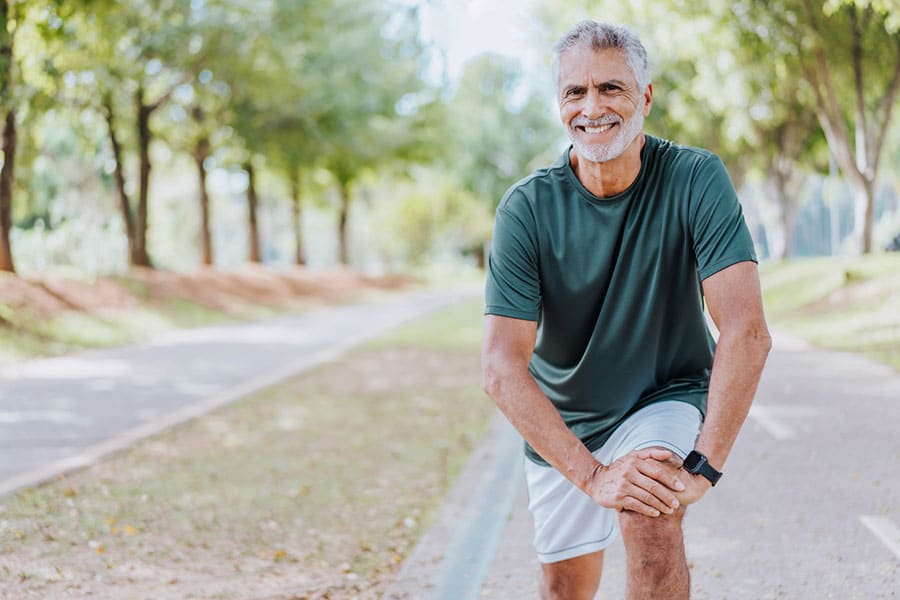 Smiling older man in athletic wear stretches outdoors on a tree-lined path, resting his hands on one knee to help relieve low back pain, with greenery and sunlight in the background.