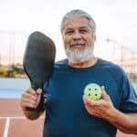 Older man with gray hair and beard, living with Degenerative Disc Disease, holding a pickleball paddle and a ball, standing on an outdoor court in Macomb County, smiling at the camera.