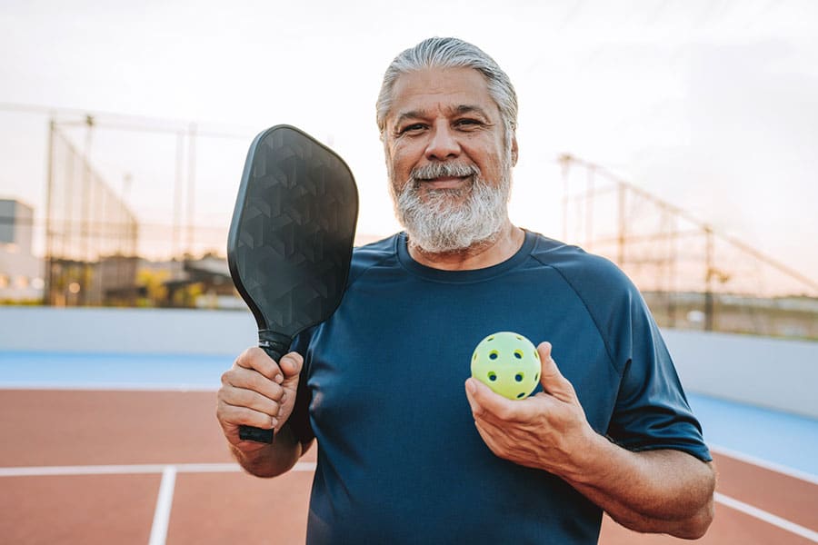 Older man with gray hair and beard, living with Degenerative Disc Disease, holding a pickleball paddle and a ball, standing on an outdoor court in Macomb County, smiling at the camera.