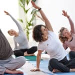 A group of adults in Macomb County sits on yoga mats indoors, stretching with one arm raised and leaning to the side. They appear focused and relaxed, engaging in gentle movements that can benefit those managing concerns like a herniated disc or degenerative disc disease. Lush plants fill the background, adding to the tranquil atmosphere.