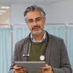 A doctor with gray hair and glasses, wearing a stethoscope and holding a tablet, stands in front of light blue privacy curtains in a medical setting, ready to discuss neuropathy symptoms and the latest options for neuropathy treatment.