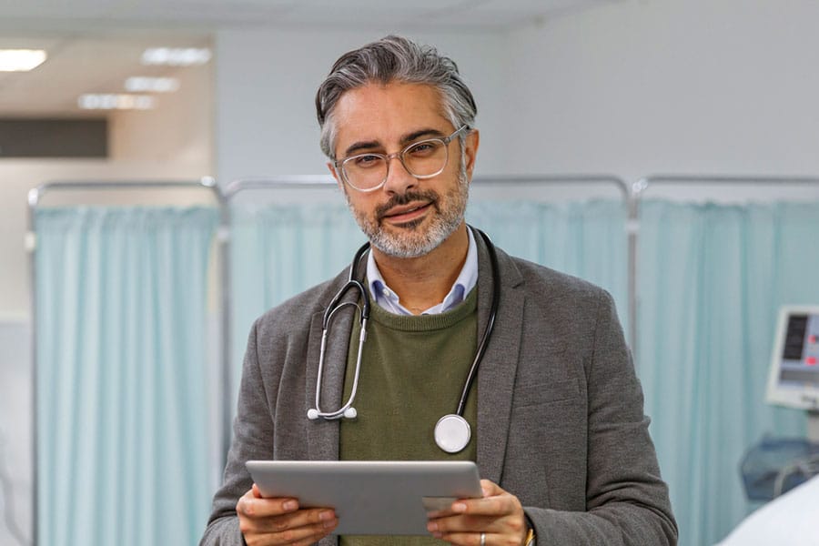 A doctor with gray hair and glasses, wearing a stethoscope and holding a tablet, stands in front of light blue privacy curtains in a medical setting, ready to discuss neuropathy symptoms and the latest options for neuropathy treatment.