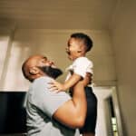 A man lifts a smiling young boy in the air inside a sunlit room, both laughing and making eye contact. Art hangs on the walls around them, embodying the joy that can follow effective treatment options—such as minimally invasive lumbar decompression—for conditions like spinal stenosis.