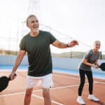 Two adults holding paddles and smiling while playing pickleball on an outdoor court on a sunny day, enjoying the benefits of pain relief that allow them to stay active.