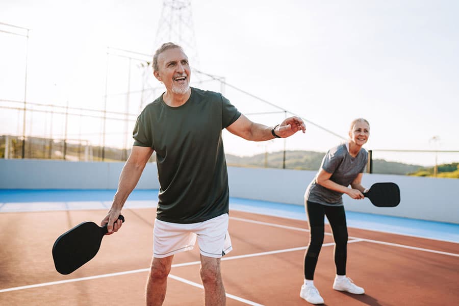 Two adults holding paddles and smiling while playing pickleball on an outdoor court on a sunny day, enjoying the benefits of pain relief that allow them to stay active.