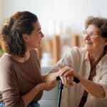 A young woman and an older woman sit on a couch, smiling at each other. The older woman holds a cane, and both appear happy and engaged in conversation about staying active and independent in a bright living room.