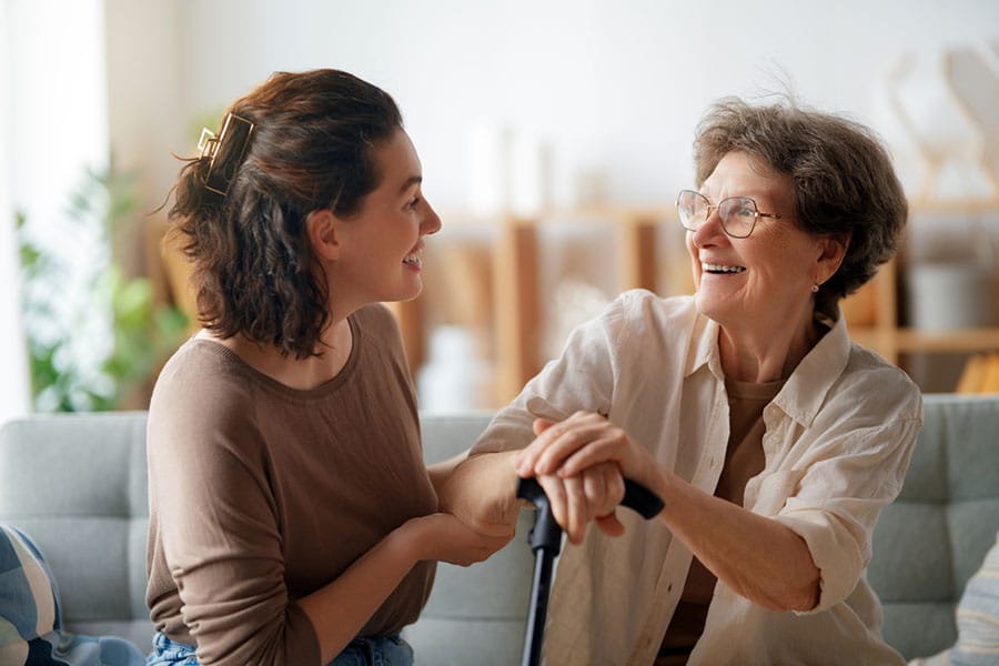 A young woman and an older woman sit on a couch, smiling at each other. The older woman holds a cane, and both appear happy and engaged in conversation about staying active and independent in a bright living room.