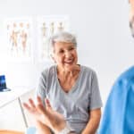 An older woman smiles while talking to a healthcare professional in a medical office, discussing effective treatments and pain management options. Anatomy posters and a laptop are visible in the background.