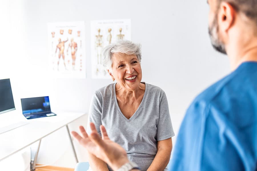 An older woman smiles while talking to a healthcare professional in a medical office, discussing effective treatments and pain management options. Anatomy posters and a laptop are visible in the background.