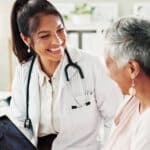 A doctor with a stethoscope smiles warmly at an older patient, who is also smiling. The doctor holds a tablet, and they appear to be having a friendly, positive conversation in a medical office as they discuss effective treatments and pain management options.