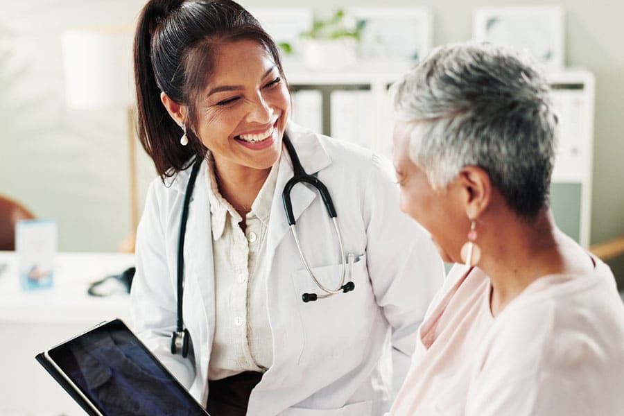A doctor with a stethoscope smiles warmly at an older patient, who is also smiling. The doctor holds a tablet, and they appear to be having a friendly, positive conversation in a medical office as they discuss effective treatments and pain management options.