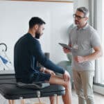 A man sits on an exam table, looking at his arm, while another man wearing glasses and holding a tablet stands nearby, discussing modern physical therapy options with him in a bright medical office.