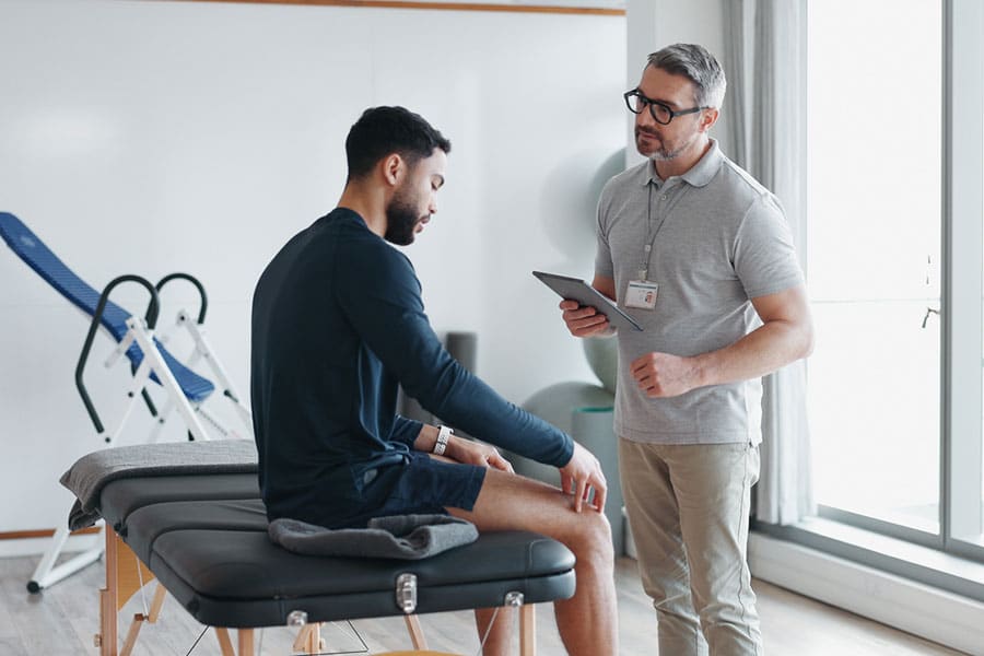 A man sits on an exam table, looking at his arm, while another man wearing glasses and holding a tablet stands nearby, discussing modern physical therapy options with him in a bright medical office.