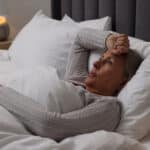A woman lies in bed under white blankets, resting her head on a pillow with one hand on her forehead, looking concerned or unwell—perhaps contemplating effective treatments for pain management as a bedside lamp glows softly in the background.