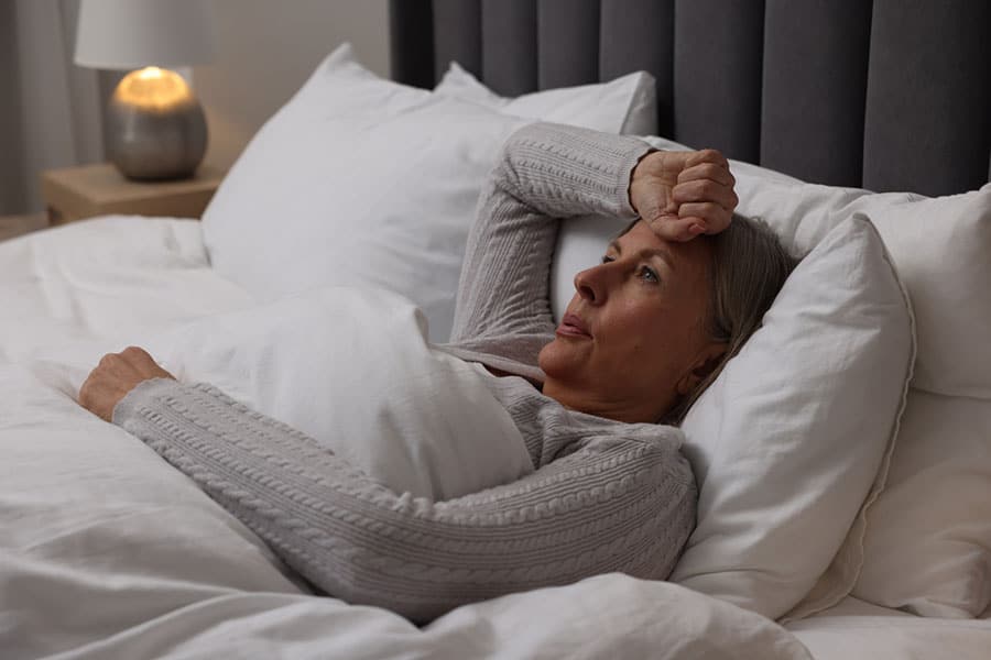 A woman lies in bed under white blankets, resting her head on a pillow with one hand on her forehead, looking concerned or unwell—perhaps contemplating effective treatments for pain management as a bedside lamp glows softly in the background.